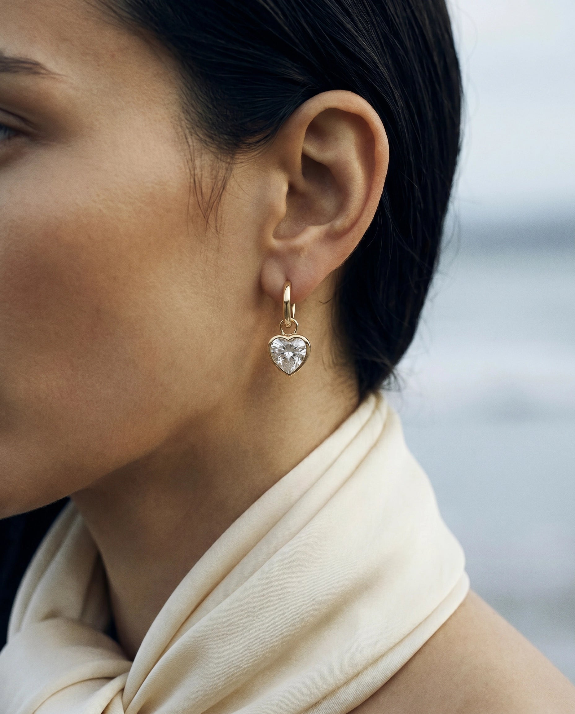 The Gold Big Love Drops — gold bezel-set heart drop earrings worn on a gold huggie hoop, editorial outdoor portrait