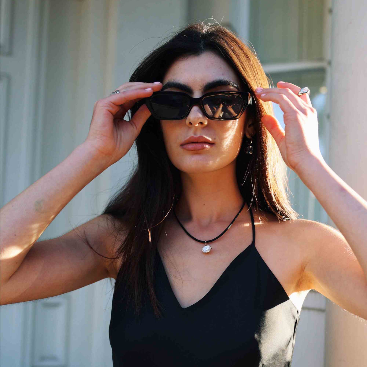 A woman wearing black cat-eye sunglasses and a black spaghetti-strap top, holding the sunglasses up to her eyes. She is wearing a black cord necklace with a white pearl pendant.