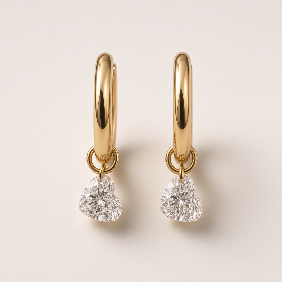 A pair of gold huggie hoop earrings with heart-shaped clear gemstones dangling from the bottom, set against a solid off-white background.